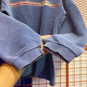 May include: A blue sweatshirt with the word "Quiksilver" printed across the chest in white and red. The sweatshirt has ribbed cuffs and a crew neck. The sleeves are long and the fabric appears to be a soft material.
