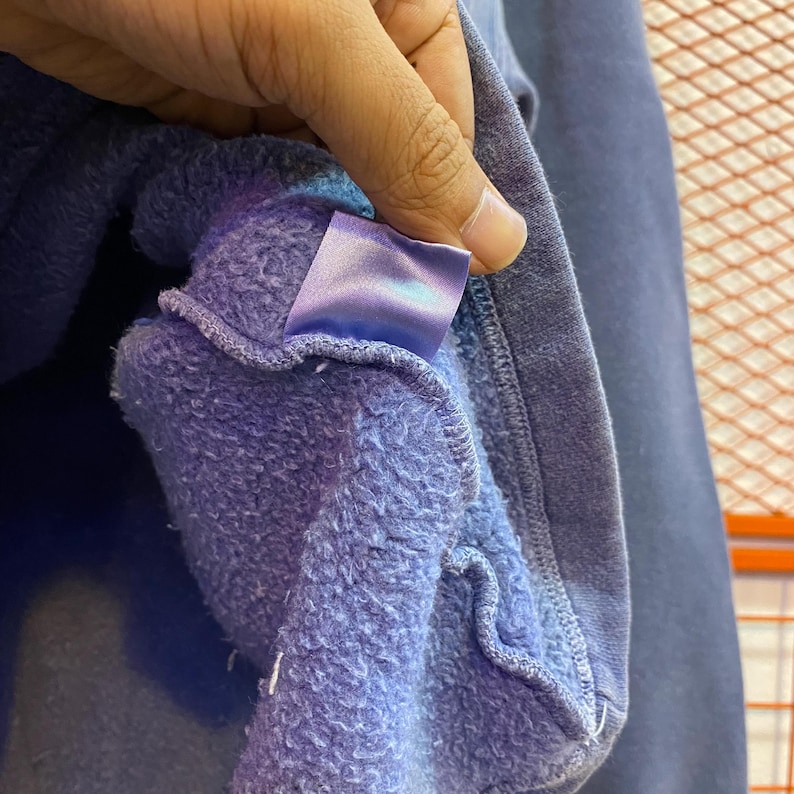 May include: Close-up of a blue sweatshirt's interior, revealing a soft, textured lining. A satin label is visible, held by a hand. The sweatshirt's exterior is a darker shade of blue, with visible stitching.