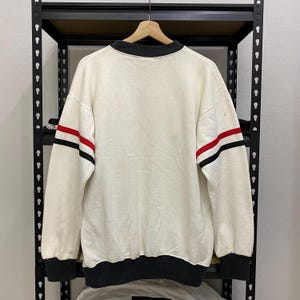 May include: Cream-colored sweatshirt with black and red stripes on the sleeves. The sweatshirt has a black collar, cuffs, and hem. The garment is hanging on a wooden hanger against a black metal shelf.