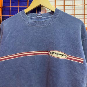 May include: A blue crew neck sweatshirt with a horizontal maroon and white stripe across the chest. The word "quiksilver" is printed in white with a red and white oval design. The sweatshirt is hanging on a wooden hanger.