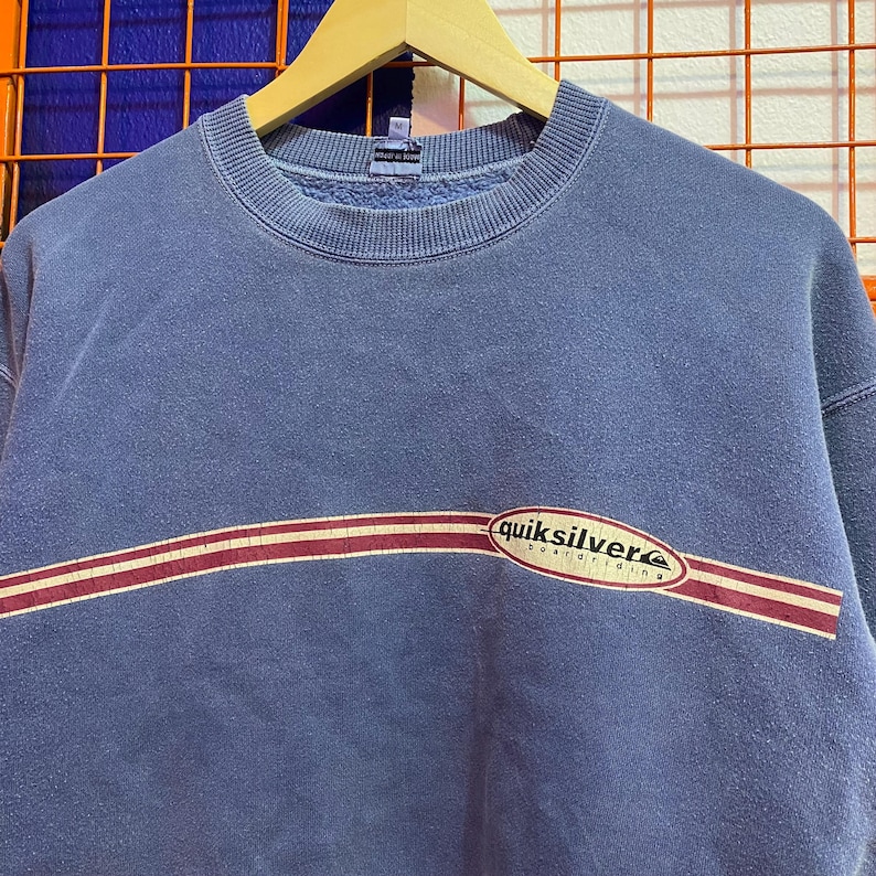 May include: A blue crew neck sweatshirt with a horizontal maroon and white stripe across the chest. The word "quiksilver" is printed in white with a red and white oval design. The sweatshirt is hanging on a wooden hanger.