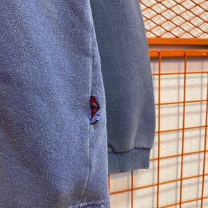 May include: Close-up of a blue sweatshirt with a small red and blue patch on the side. The sweatshirt has a ribbed cuff and a slightly worn appearance, suggesting it's been well-loved.