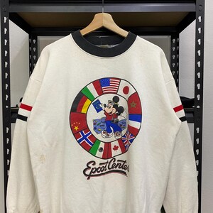 May include: Cream-colored sweatshirt with a black collar and red and black stripes on the sleeves. The front features a circular graphic of Mickey Mouse surrounded by flags and the text "Epcot Center".