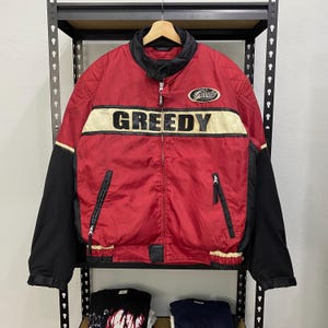 Può includere: Una giacca rossa e nera con la parola "GREEDY" in lettere bianche sul davanti. La giacca ha una chiusura a zip e un colletto nero.