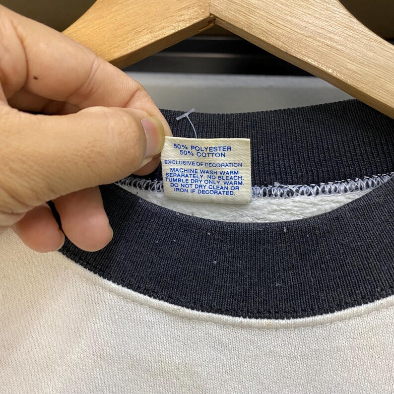 May include: Close-up of a white and black sweatshirt collar. A tag with the text "50% POLYESTER 50% COTTON" is held up. The tag also includes washing instructions: "MACHINE WASH WARM SEPARATELY. NO BLEACH. TUMBLE DRY ONLY, WARM. DO NOT DRY CLEAN OR IRON IF DECORATED."