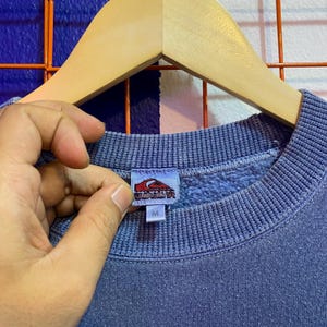 May include: A blue sweatshirt with a ribbed crew neck and cuffs. The tag features a red and white logo and the letter "M". The sweatshirt is hanging on a wooden hanger.