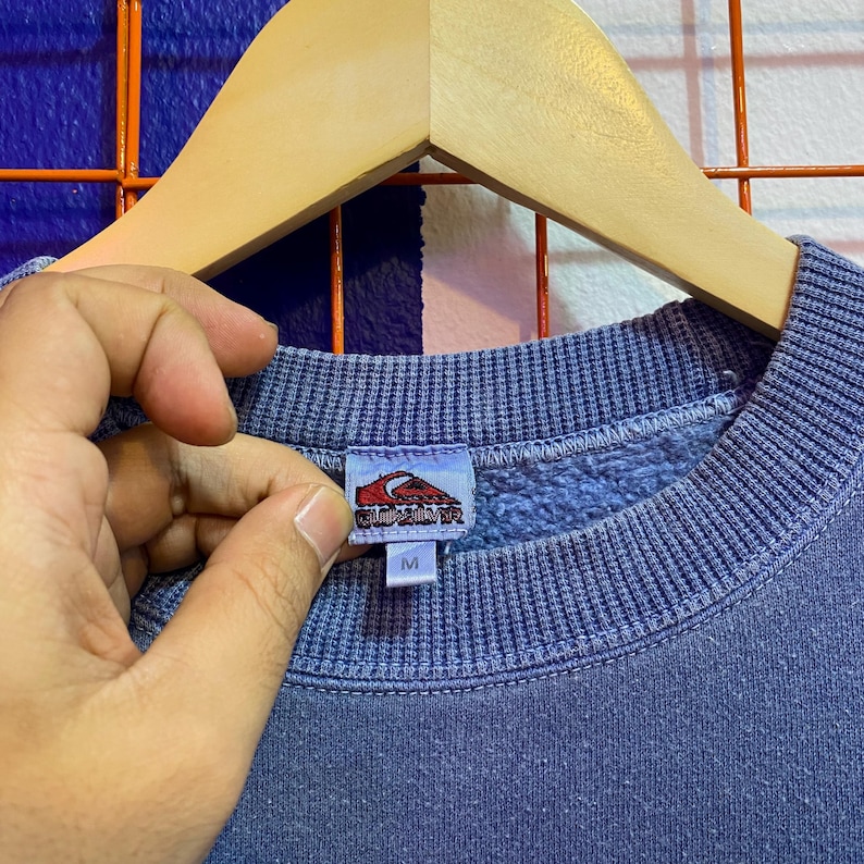 May include: A blue sweatshirt with a ribbed crew neck and cuffs. The tag features a red and white logo and the letter "M". The sweatshirt is hanging on a wooden hanger.