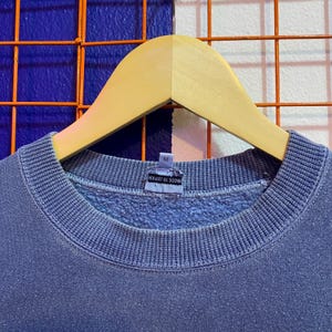 May include: A blue crew neck sweatshirt hanging on a wooden hanger. The sweatshirt has a ribbed collar and cuffs. The tag reads "MADE IN JAPAN" and has a size "M". The background is a mix of blue and white.