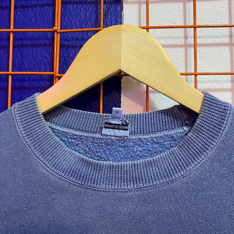 May include: A blue crew neck sweatshirt hanging on a wooden hanger. The sweatshirt has a ribbed collar and cuffs. The tag reads "MADE IN JAPAN" and has a size "M". The background is a mix of blue and white.