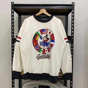 May include: Cream-colored sweatshirt featuring a circular graphic with Mickey Mouse and flags from various countries. The sweatshirt has black and red stripes on the sleeves and the text "Epcot Center" below the graphic. The collar and cuffs are black.