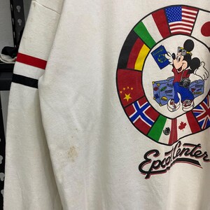 May include: Off-white sweatshirt featuring a colorful graphic of Mickey Mouse as a traveler, surrounded by flags of different countries. Mickey holds a passport and stands next to a suitcase. The word "Epcot Center" is written in script below the graphic.
