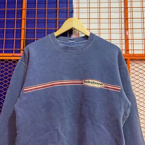 May include: A faded blue sweatshirt with a crew neck and long sleeves. The sweatshirt features a horizontal stripe design in red and white across the chest, with the "quiksilver" logo in white and green.