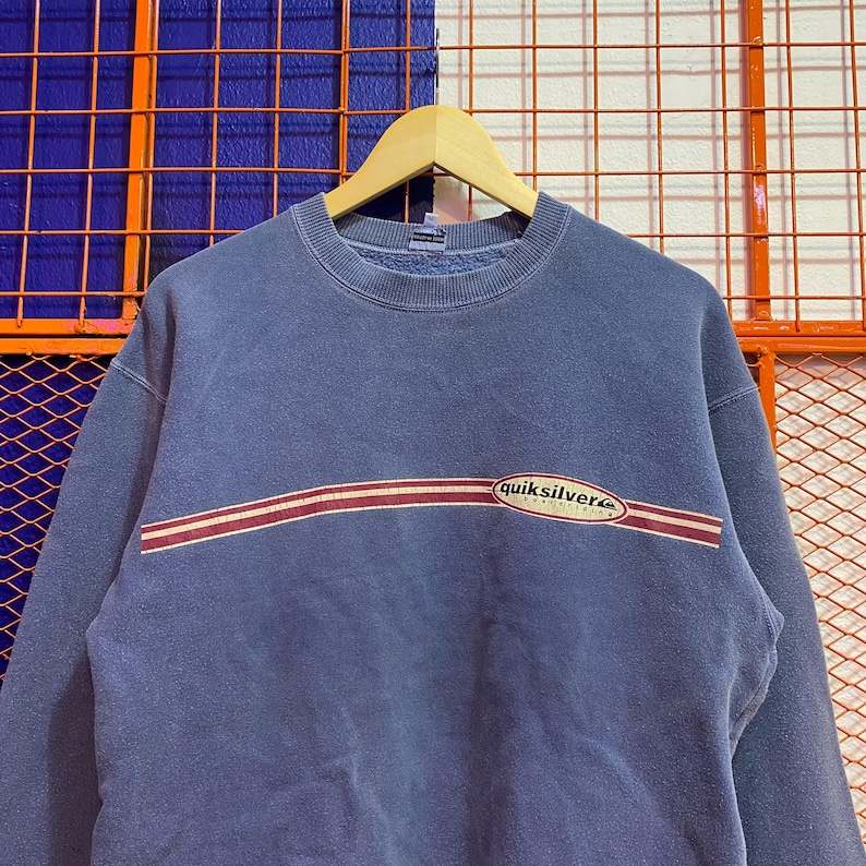 May include: A faded blue sweatshirt with a crew neck and long sleeves. The sweatshirt features a horizontal stripe design in red and white across the chest, with the "quiksilver" logo in white and green.