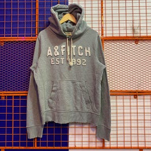 May include: A grey hoodie with the text "A&FITCH EST 1892" in white, featuring a drawstring hood and a front pocket. The hoodie is displayed on a wooden hanger against a backdrop of blue and orange wire mesh.