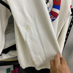 May include: Cream-colored sweatshirt with black accents and a graphic featuring a cartoon character and flags. The sleeve shows some discoloration. The sweatshirt is made of a soft material.