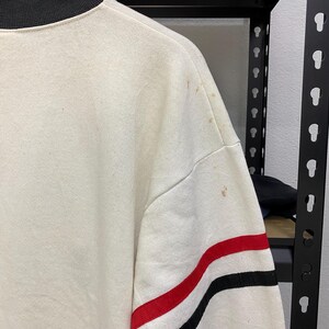 May include: Cream-colored sweatshirt with a black collar and red and black stripes on the sleeve. The sweatshirt has some brown stains on the shoulder. The garment is hanging on a rack.