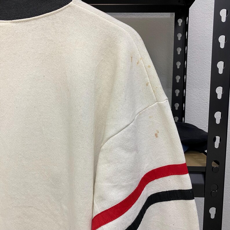 May include: Cream-colored sweatshirt with a black collar and red and black stripes on the sleeve. The sweatshirt has some brown stains on the shoulder. The garment is hanging on a rack.