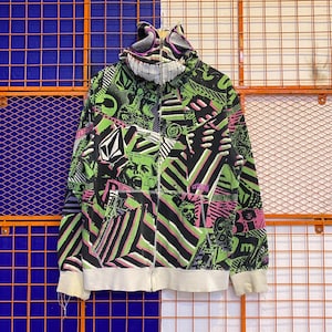 May include: A black, green, and pink zip-up hoodie with a unique design. The hoodie features a graphic pattern, a hood with a smiling face design, and white cuffs and hem. The Volcom logo is visible on the front.