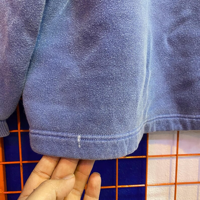 May include: Close-up of a faded periwinkle blue sweatshirt. The bottom hem is visible, with a ribbed band and a small white mark. The fabric has a slightly worn, textured appearance, suggesting it's a well-loved garment.