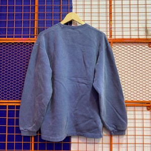 May include: A faded blue sweatshirt with a crew neck and long sleeves. The garment is hanging on a wooden hanger. The sweatshirt has a vintage, worn appearance. The background features a grid pattern in orange and blue.