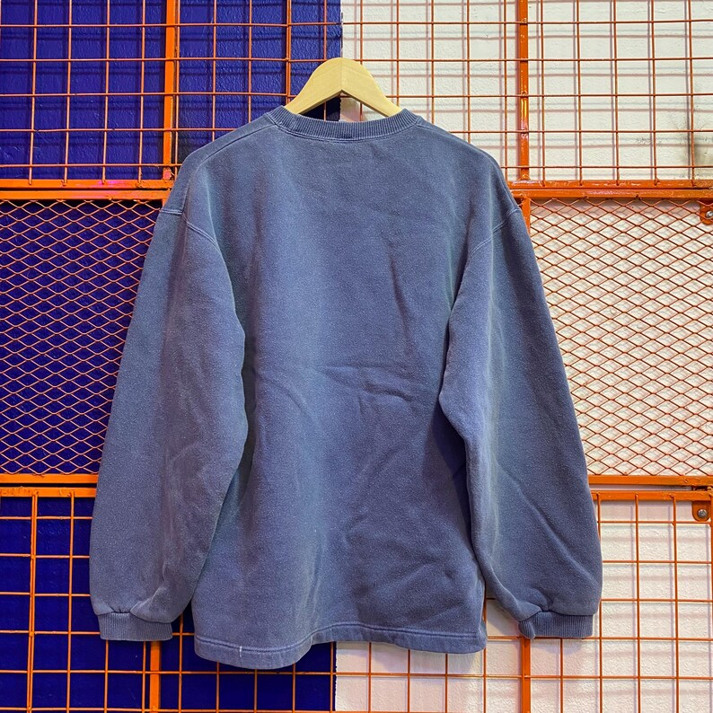 May include: A faded blue sweatshirt with a crew neck and long sleeves. The garment is hanging on a wooden hanger. The sweatshirt has a vintage, worn appearance. The background features a grid pattern in orange and blue.