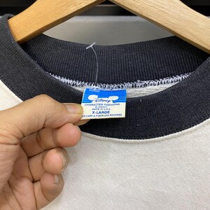 May include: Close-up of a vintage Disney sweatshirt. The sweatshirt has a white body with black sleeves and collar. A blue and white tag reads "Disney CHARACTER FASHIONS MADE IN U.S.A. X-LARGE". The tag is held by a hand.