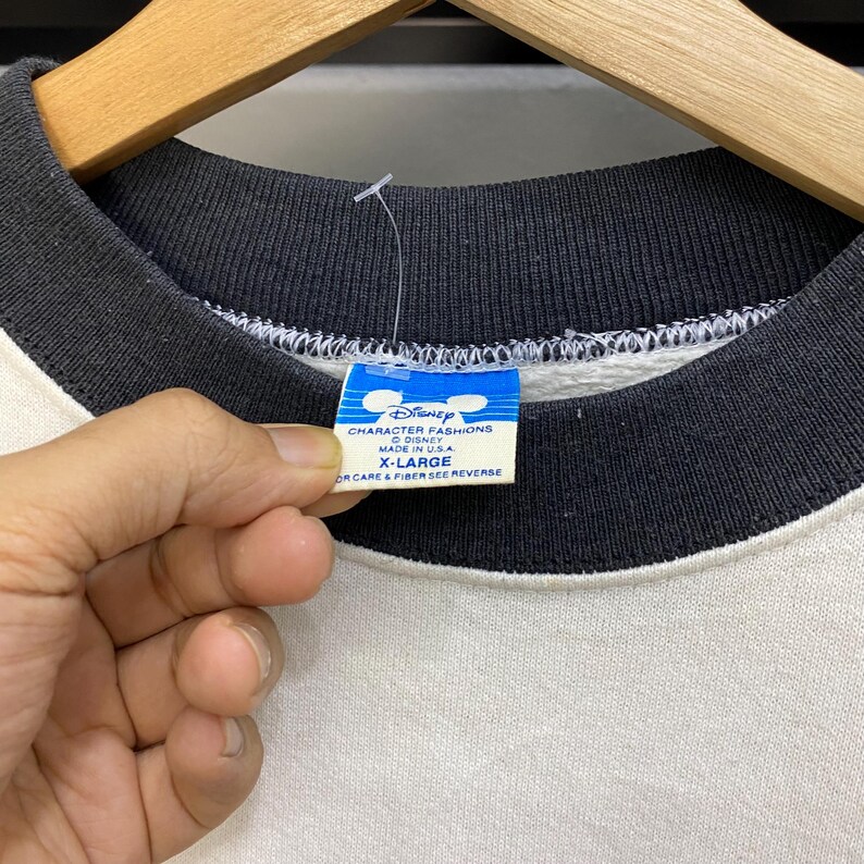 May include: Close-up of a vintage Disney sweatshirt. The sweatshirt has a white body with black sleeves and collar. A blue and white tag reads "Disney CHARACTER FASHIONS MADE IN U.S.A. X-LARGE". The tag is held by a hand.