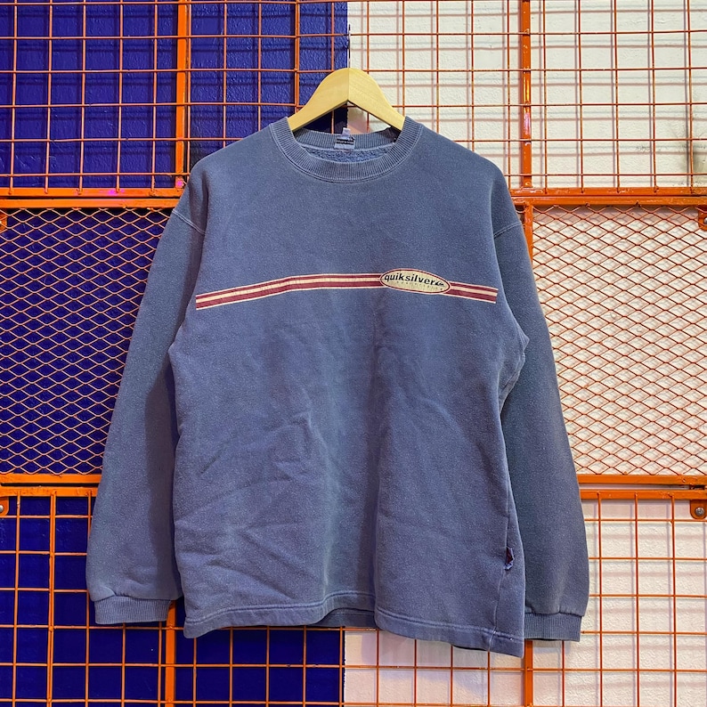May include: A faded blue sweatshirt with a crew neck and long sleeves. The shirt features a horizontal red and white stripe across the chest with the brand name "Quiksilver" in a white oval. The sweatshirt is hanging on a wooden hanger.
