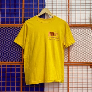 May include: A yellow short-sleeved t-shirt with the text "Holley Carburetor Co. Detroit, Michigan" printed in red. The shirt is hanging on a wooden hanger against a grid background.