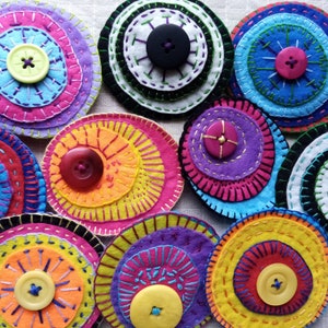 May include: A collection of colourful felt circles with embroidered designs and buttons. Each circle features multiple layers of felt in various colours, creating a layered and textured effect. The buttons are a mix of colours and sizes, adding to the overall visual appeal.