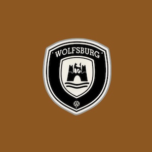 以下が含まれることがあります： 黒と白のエンブレムで、円の中に「WOLFSBURG」という文字が書かれており、その周りに狼が乗ったスタイリッシュな城が描かれています。エンブレムは茶色の背景です。
