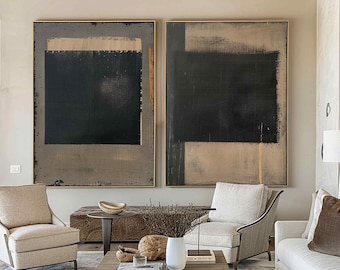 May include: Two large abstract paintings with black and tan brushstrokes hang above a living room with neutral-toned furniture. The room includes two beige armchairs, a wooden coffee table, and a white sofa. The ceiling has exposed wooden beams.