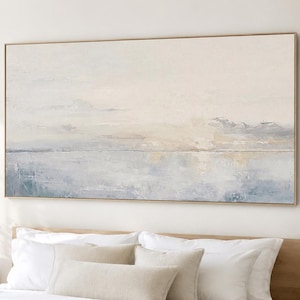 Puede incluir: Una gran pintura abstracta rectangular en un marco dorado, que representa un paisaje sereno con suaves tonos azules, blancos y beige. La pintura cuelga sobre una cama con sábanas blancas y almohadas beige.