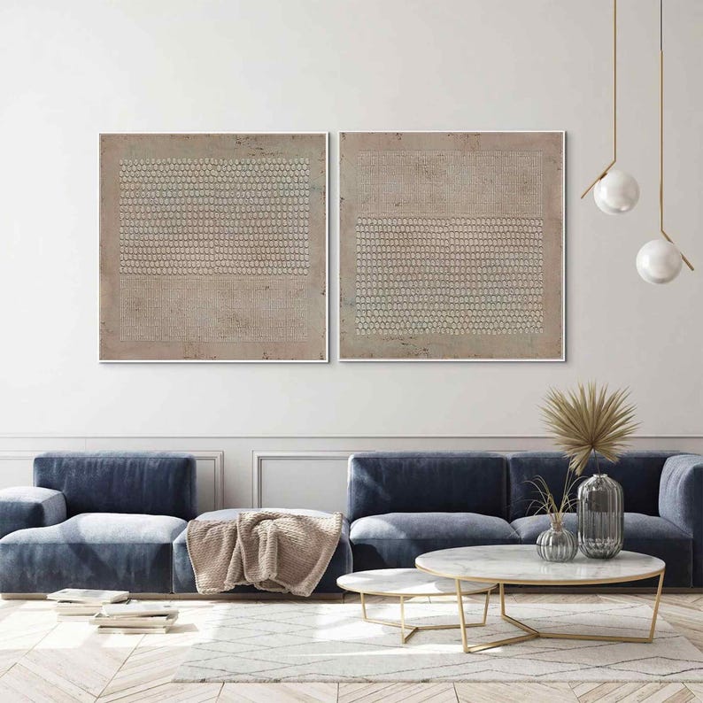 Puede incluir: Dos obras de arte abstractas enmarcadas con una paleta de colores neutros. Cada pieza presenta un dise&ntilde;o texturizado con patrones repetidos en beige y blanco roto, creando una est&eacute;tica minimalista. La obra de arte se exhibe sobre un sof&aacute; azul en un sal&oacute;n moderno.
