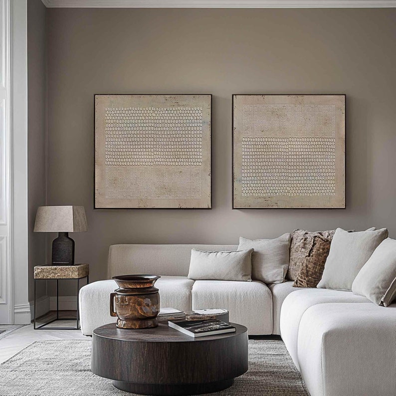 Puede incluir: Escena de sal&oacute;n con est&eacute;tica moderna. Dos grandes obras de arte abstractas cuadradas cuelgan de la pared. Un sof&aacute; seccional blanco, una mesa de centro de madera oscura y una mesa auxiliar con una l&aacute;mpara completan el espacio. Los tonos neutros dominan la decoraci&oacute;n.