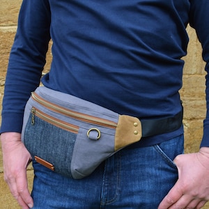 May include: A gray and blue canvas fanny pack with a brown leather flap and a black adjustable strap. The fanny pack has two zippered pockets and a small pocket on the front.