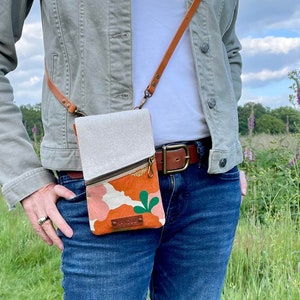 May include: A crossbody phone bag with a tan canvas top and a patterned orange and pink bottom. The bag has a brown leather strap and a gold zipper. The bag is worn over a light green jacket and blue jeans.