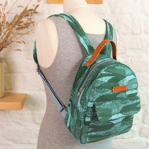 May include: A small green and blue patterned backpack with a brown leather handle and adjustable straps. The backpack has a zipper closure and a front pocket.