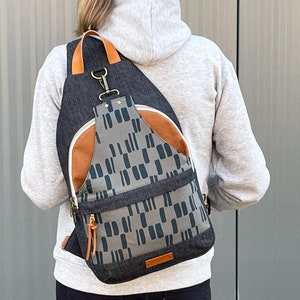 May include: A gray and black canvas backpack with a brown leather flap and a geometric pattern. The backpack has a single adjustable strap and a zipper closure.