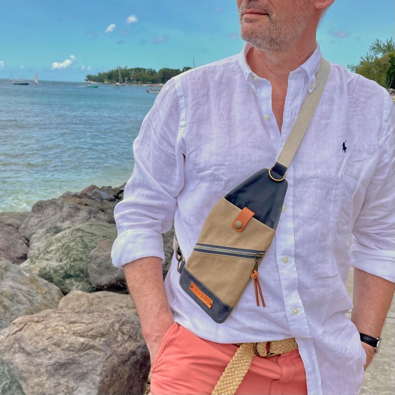 K&ouml;nnte beinhalten: Eine Person tr&auml;gt ein wei&szlig;es Leinenhemd mit einer braunen und schwarzen Umh&auml;ngetasche. Die Tasche hat einen Rei&szlig;verschluss und einen kleinen Lederriemen. Die Person steht an einem Strand mit Felsen und Wasser im Hintergrund.