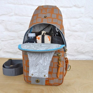 May include: A brown and gray patterned sling bag with a blue zipper and a gray strap. The bag is open and has a water bottle, a small pouch, and a black item inside.