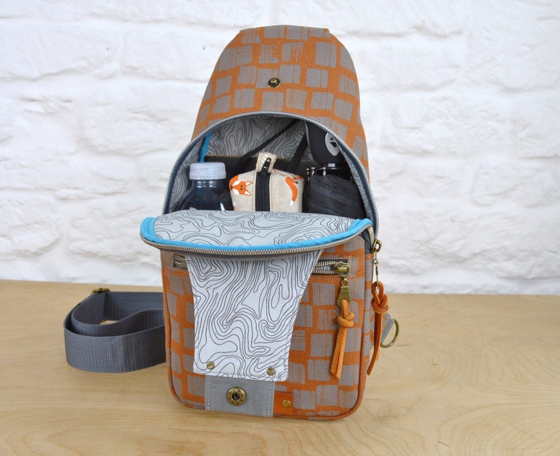 May include: A brown and gray patterned sling bag with a blue zipper and a gray strap. The bag is open and has a water bottle, a small pouch, and a black item inside.