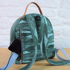 May include: A green and blue patterned backpack with a brown leather handle and a zipped pocket on the front. A black phone is in the pocket.