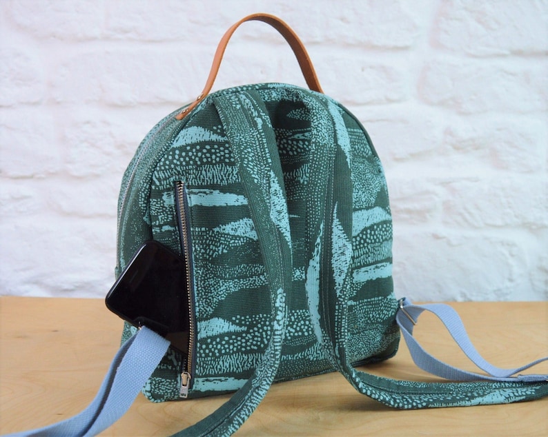 May include: A green and blue patterned backpack with a brown leather handle and a zipped pocket on the front. A black phone is in the pocket.