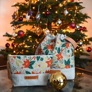 Pu&ograve; includere: Due borse in tessuto con stampa floreale natalizia in rosso, verde e oro. Le borse sono in lino e hanno una chiusura a coulisse. La borsa pi&ugrave; piccola ha la scritta "KANDOU" stampata sul davanti. Le borse sono appoggiate su un tavolo davanti a un albero di Natale.