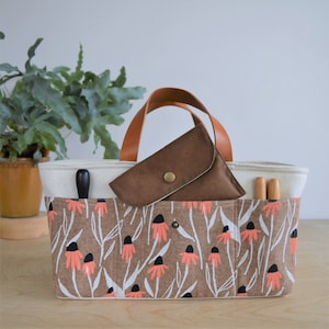 May include: A rectangular tote bag with a floral print in brown, white, and coral. The bag has a cream-colored interior and brown leather handles. A small brown pouch with a button closure sits inside the bag, along with two wooden tools.