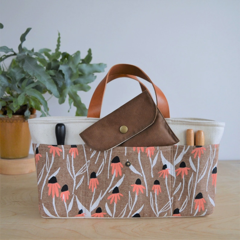 May include: A rectangular tote bag with a floral print in brown, white, and coral. The bag has a cream-colored interior and brown leather handles. A small brown pouch with a button closure sits inside the bag, along with two wooden tools.