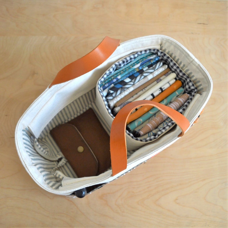 May include: Cream-colored tote bag with brown leather handles and accents. The bag has a striped interior and contains a brown leather wallet and a compartment filled with colorful fabric rolls. The bag is open, revealing its contents.