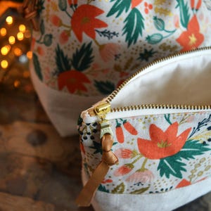 Pu&ograve; includere: Una pochette bianca con cerniera con stampa floreale con fiori rossi, rosa e verdi. La pochette ha una cerniera dorata e un tiretto in pelle marrone.