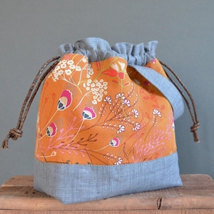 May include: A drawstring bag with a floral print in shades of orange, pink, and white. The bag has a gray linen lining and brown drawstrings.